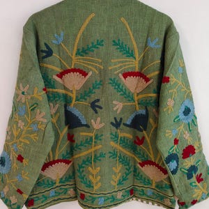May include: A green jacket with intricate floral embroidery in red, blue, yellow, and beige. The jacket features a detailed design on the back and sleeves, with a textured fabric. The embroidery includes flowers and leaves, creating a decorative outerwear piece.
