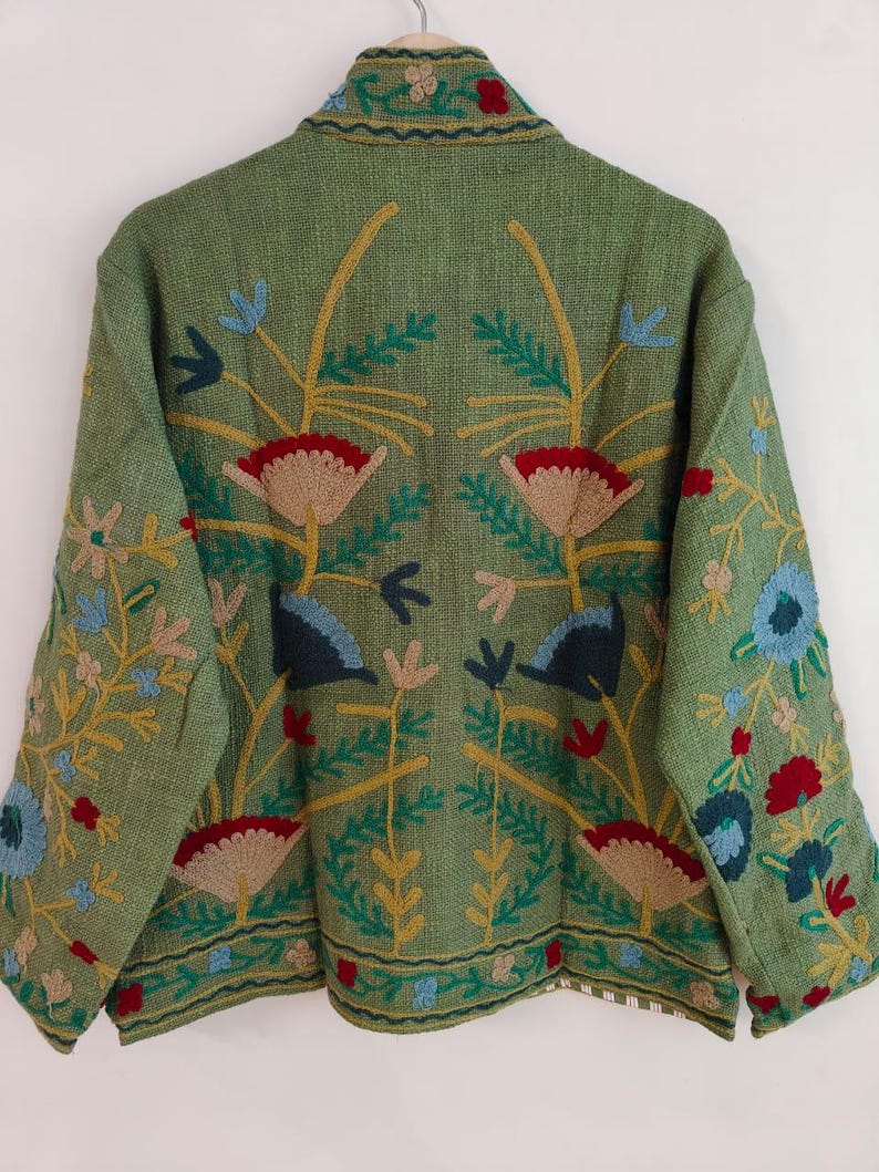 May include: A green jacket with intricate floral embroidery in red, blue, yellow, and beige. The jacket features a detailed design on the back and sleeves, with a textured fabric. The embroidery includes flowers and leaves, creating a decorative outerwear piece.
