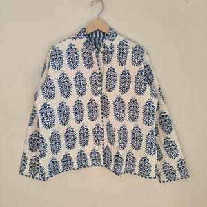 May include: A reversible jacket with a blue and white floral pattern. The jacket has a button-up front and a contrasting blue striped lining. It is hanging on a metal hanger against a plain white wall.