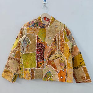 Può includere: Giacca patchwork nei toni del giallo, arancione e beige, con collo alla coreana. La giacca è realizzata con vari pezzi di tessuto, alcuni con dettagli ricamati e piccoli specchi. È appesa a un'appendiabiti di legno.
