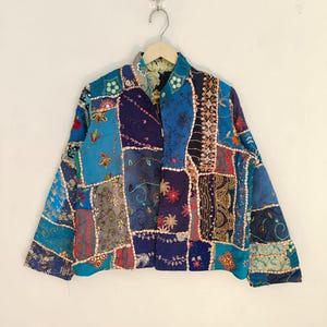 Puede incluir: Chaqueta patchwork en tonos azules variados, con bordados florales y geométricos. La chaqueta está hecha de diferentes cuadrados de tela, creando un diseño único. Cuelga de una percha de madera sobre un fondo blanco.