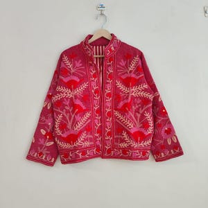 May include: A magenta jacket with intricate floral embroidery in shades of red and pink. The jacket features a high collar, long sleeves, and a striped lining. The floral design is detailed and covers the front, sleeves, and collar.