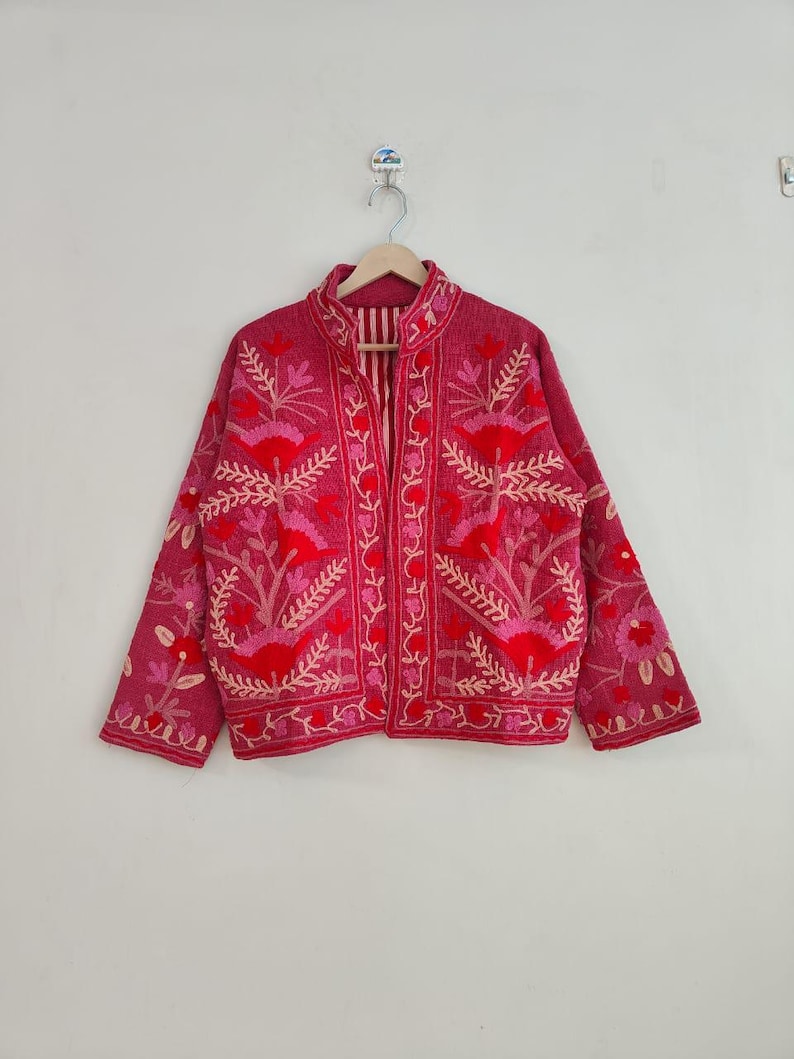 May include: A magenta jacket with intricate floral embroidery in shades of red and pink. The jacket features a high collar, long sleeves, and a striped lining. The floral design is detailed and covers the front, sleeves, and collar.