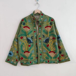 May include: A green embroidered jacket with a stand-up collar. The jacket features a floral design in red, blue, yellow, and tan. The jacket is hanging on a wooden hanger against a white background.