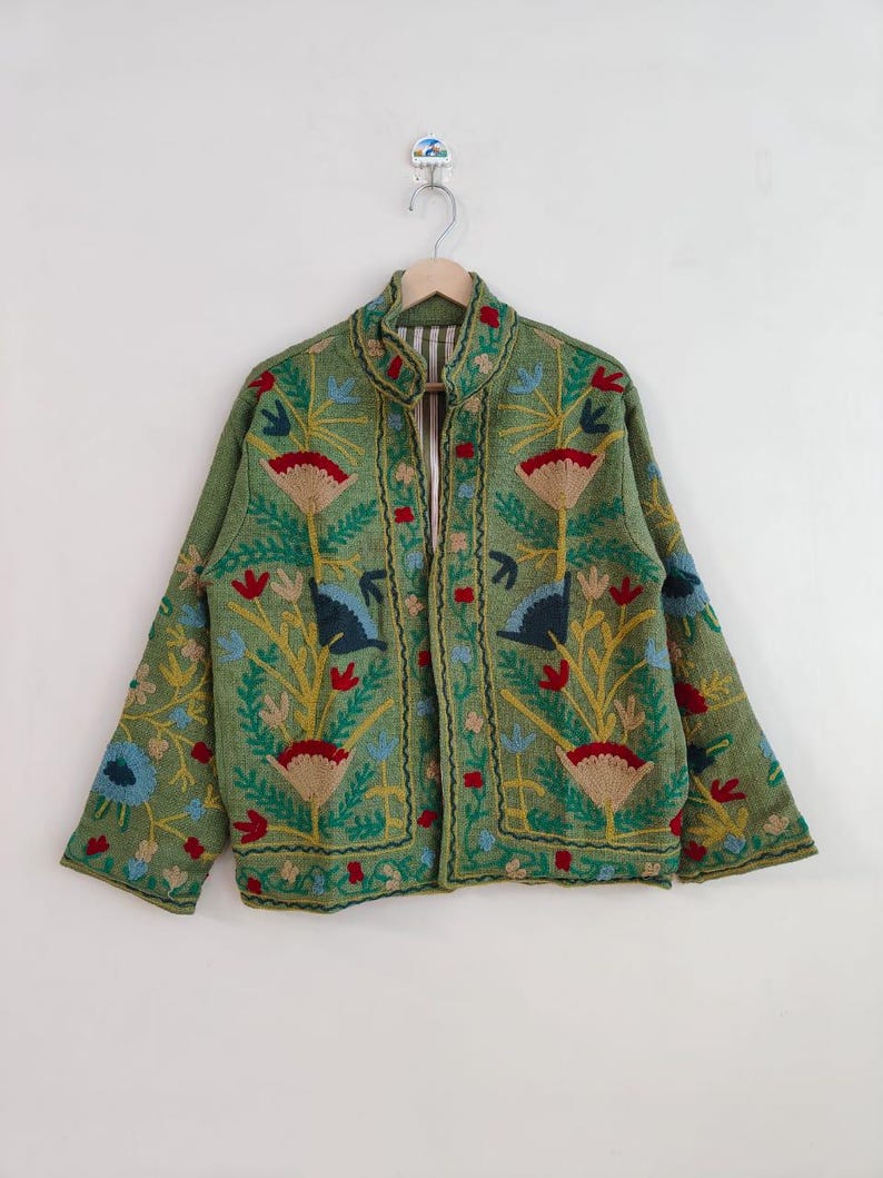 May include: A green embroidered jacket with a stand-up collar. The jacket features a floral design in red, blue, yellow, and tan. The jacket is hanging on a wooden hanger against a white background.