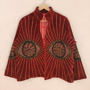 Pode incluir: Casaco de veludo bordô com gola alta e mangas compridas. O casaco apresenta um grande design de olho na frente, com linhas radiantes. O forro interior é listrado em vermelho e branco. O casaco está pendurado em um cabide de madeira.