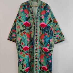 Op de afbeelding: Een lange, open jas in een lichtgroene tint, versierd met levendige bloemenborduurwerk. Het ontwerp toont grote roze en rode bloemen, turquoise bladeren en blauwe accenten. De jas hangt aan een houten hanger tegen een effen witte achtergrond.