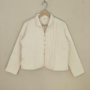May include: Off-white quilted jacket with a mandarin collar and long sleeves. The jacket has a button-up front and a subtle quilted design. It is suspended on a wooden hanger against a plain background.