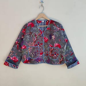 May include: A grey quilted jacket with a floral pattern of red and purple flowers, green leaves, and birds. The jacket has two front pockets and is trimmed with blue and white striped fabric. The jacket is hanging on a wooden hanger.