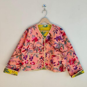 May include: A reversible jacket with a floral design in pink, red, purple, and green. The jacket has a quilted texture, two front pockets, and yellow trim. The sleeves are long, and the jacket is hanging on a wooden hanger.