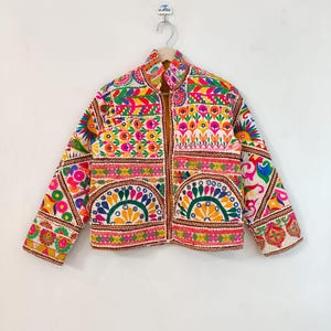 Puede incluir: Chaqueta bordada colorida con cuello alto. La chaqueta presenta una vibrante mezcla de patrones florales y geométricos en tonos rosa, amarillo, verde y azul. La chaqueta está colgada en una percha de madera sobre un fondo blanco.