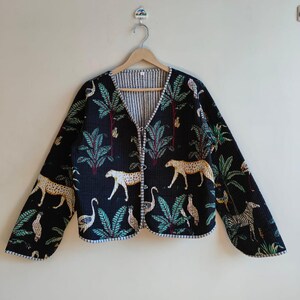May include: Black quilted jacket with a V-neck and long sleeves. The jacket features a repeating pattern of leopards, birds, and palm trees in shades of tan, green, and white. The jacket is hanging on a wooden hanger.