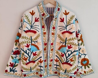May include: Cream-colored embroidered jacket with a mandarin collar. The jacket features colorful floral embroidery in red, orange, blue, and green. The embroidery is detailed and covers the front, sleeves, and collar of the jacket. The jacket is hanging on a wooden hanger.