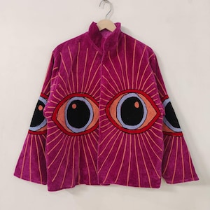 Op de afbeelding: Een magenta fluwelen jas met een hoge kraag en lange mouwen. De jas heeft grote geborduurde oogontwerpen op de voorkant en mouwen, met oranje en zwarte details. Het ontwerp bevat stralende lijnen.