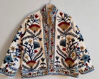 May include: Cream-colored jacket with intricate floral embroidery in red, blue, and brown. The jacket features a stand-up collar and is displayed on a hanger against a plain white wall. The embroidery creates a detailed, textured pattern.
