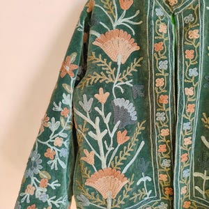 Puede incluir: Chaqueta de terciopelo verde con intrincados bordados florales en tonos melocot&oacute;n, azul y crema. La chaqueta presenta un cuello mandar&iacute;n y bordados detallados en las mangas y bordes delanteros, de estilo bohemio.