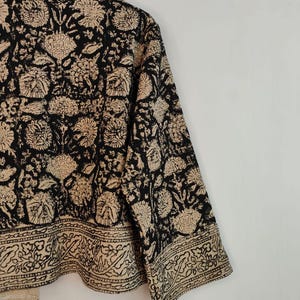K&ouml;nnte beinhalten: Eine schwarz-beige Jacke mit floralem Muster. Die Jacke hat ein sich wiederholendes Blumenmuster in Beige auf schwarzem Hintergrund. Die &Auml;rmel sind dreiviertellang und die Jacke h&auml;ngt an einem Holzb&uuml;gel.