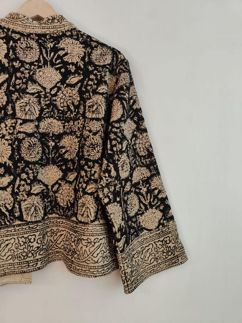 K&ouml;nnte beinhalten: Eine schwarz-beige Jacke mit floralem Muster. Die Jacke hat ein sich wiederholendes Blumenmuster in Beige auf schwarzem Hintergrund. Die &Auml;rmel sind dreiviertellang und die Jacke h&auml;ngt an einem Holzb&uuml;gel.
