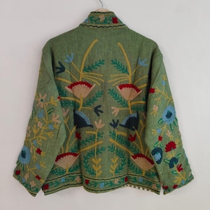 May include: A green jacket with floral embroidery. The jacket features a high collar and long sleeves. The embroidery includes red, blue, yellow, and beige flowers and leaves. The jacket is hanging on a metal hanger against a white wall.