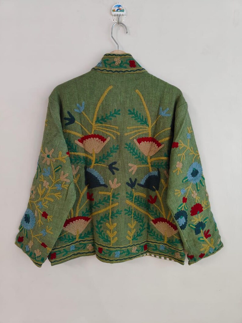 May include: A green jacket with floral embroidery. The jacket features a high collar and long sleeves. The embroidery includes red, blue, yellow, and beige flowers and leaves. The jacket is hanging on a metal hanger against a white wall.