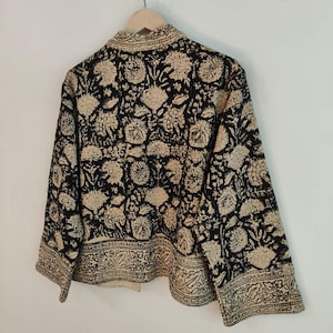 K&ouml;nnte beinhalten: Schwarze Jacke mit beigem Blumenmuster. Die Jacke h&auml;ngt an einem Holzb&uuml;gel. Das florale Design bedeckt die gesamte Jacke, einschlie&szlig;lich Kragen und Manschetten. Die Jacke hat einen dekorativen Rand.
