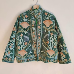 Puede incluir: Chaqueta de terciopelo verde con intrincados bordados florales en tonos melocot&oacute;n, azul y blanco. La chaqueta, colgada de una percha de madera, presenta un cuello mandar&iacute;n. El bordado cubre el frente, las mangas y el cuello.