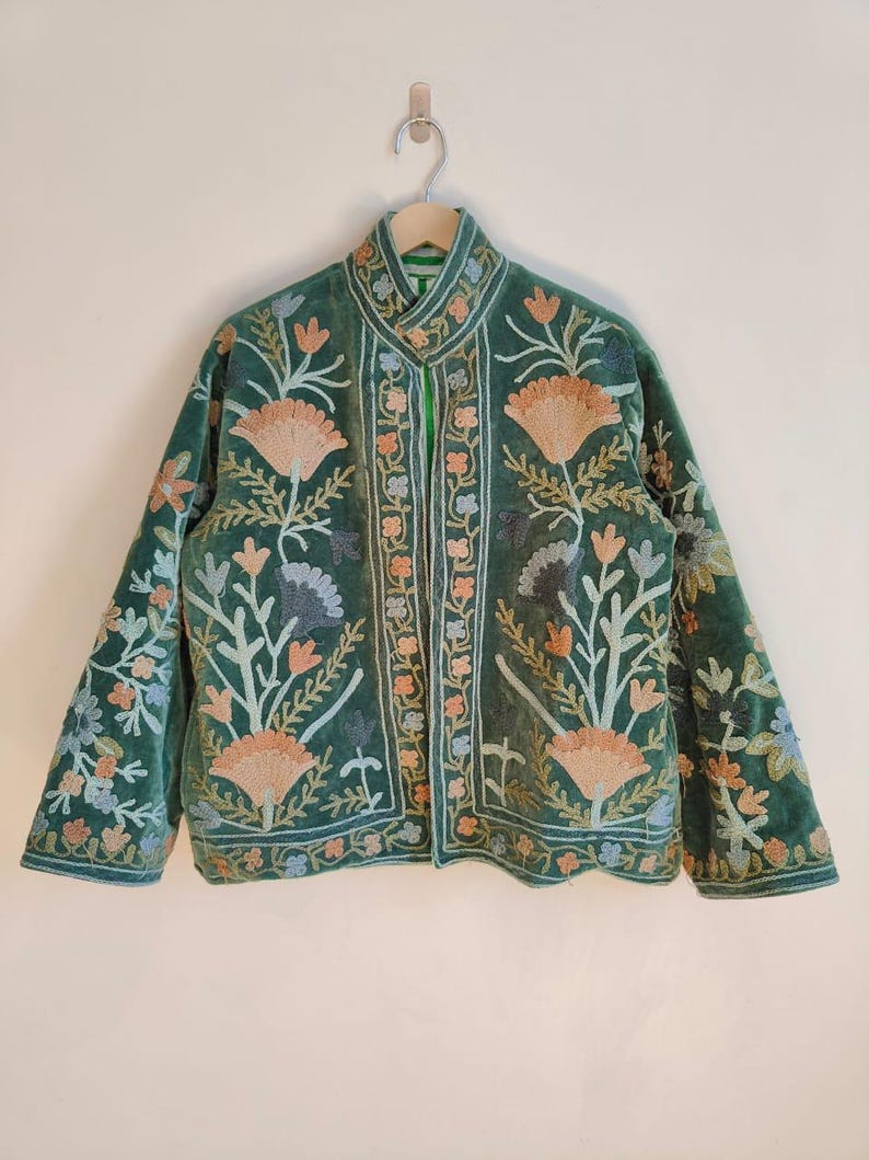 Puede incluir: Chaqueta de terciopelo verde con intrincados bordados florales en tonos melocot&oacute;n, azul y blanco. La chaqueta, colgada de una percha de madera, presenta un cuello mandar&iacute;n. El bordado cubre el frente, las mangas y el cuello.