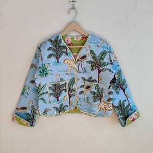 May include: A light blue jacket with a tropical print featuring palm trees, leopards, and birds. The jacket has a green floral lining and trim, with two front pockets. The jacket is hanging on a wooden hanger.