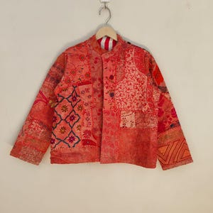 Puede incluir: Chaqueta patchwork en tonos rojos, naranjas y rosas, con varios diseños florales y geométricos. La chaqueta tiene cuello mandarín y cierres de botón. Las mangas son largas y la tela parece una mezcla de texturas.