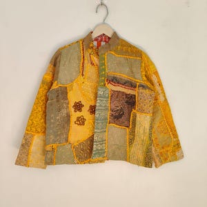 May include: A patchwork jacket in shades of yellow, olive green, and brown. The jacket features a mandarin collar and is composed of various fabric squares with floral and geometric patterns. The jacket is hanging on a wooden hanger.