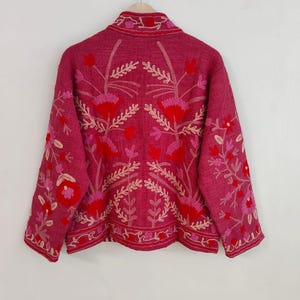 May include: A magenta jacket with intricate floral embroidery in shades of red and pink. The jacket features a collared neckline and long sleeves, with the floral design covering the back and sleeves. The jacket is hanging on a hanger against a white background.