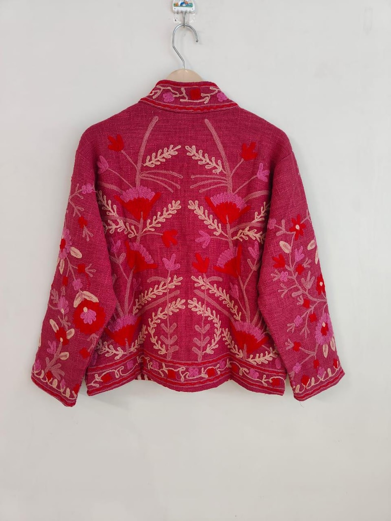 May include: A magenta jacket with intricate floral embroidery in shades of red and pink. The jacket features a collared neckline and long sleeves, with the floral design covering the back and sleeves. The jacket is hanging on a hanger against a white background.