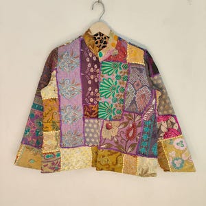 Op de afbeelding: Een patchwork jasje met diverse bloemen- en paisley patronen in paars, goud, groen en blauw. De jas hangt aan een houten hanger.