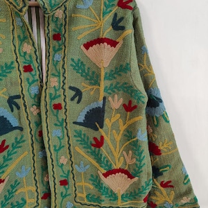 May include: A green jacket with intricate floral embroidery in red, blue, yellow, and tan. The jacket features a collar and is lined with a striped fabric. The embroidery creates a textured, detailed design.