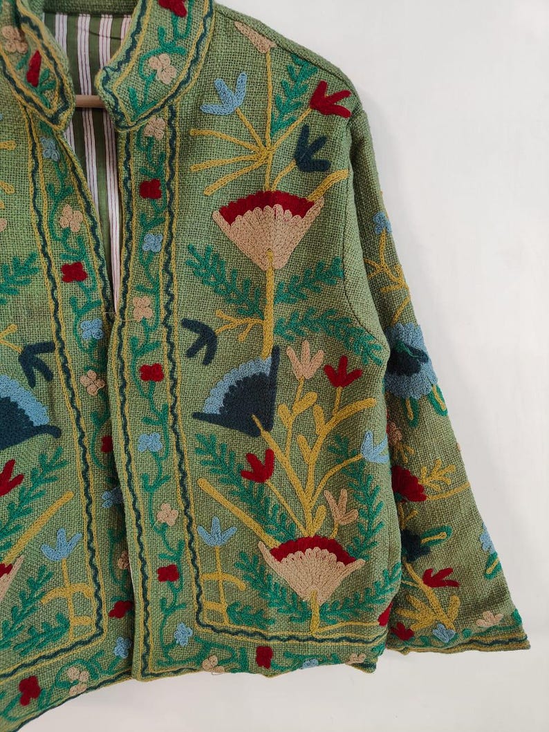 May include: A green jacket with intricate floral embroidery in red, blue, yellow, and tan. The jacket features a collar and is lined with a striped fabric. The embroidery creates a textured, detailed design.