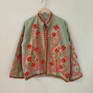 May include: Sage green jacket with red and pink floral embroidery. The jacket features a mandarin collar and a striped lining visible at the hem and collar. The embroidery creates a symmetrical design on the front and sleeves.