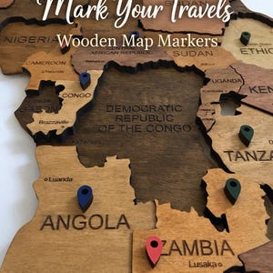 May include: Wooden map of Africa with country names and colorful pin markers. The text "Mark Your Travels Wooden Map Markers" is displayed across the top. The map is made of wood with varying shades of brown.