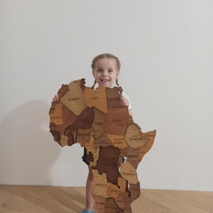 May include: A wooden map of Africa with country names engraved. The map is made of various shades of brown wood. A young person is holding the map. The map has the words "WOW MAP" in the bottom right corner.