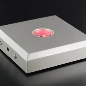 Op de afbeelding: Een vierkante, zilverkleurige displaybasis met een rond, rood LED-licht in het midden.