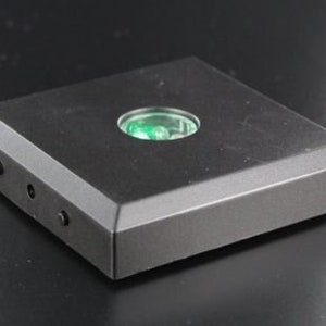 Könnte beinhalten: Ein schwarzer, quadratischer Präsentationssockel mit abgeschrägter Kante. Er hat einen kreisförmigen, transparenten Einsatz mit grünem Licht. Drei kleine, dunkle Schrauben sind an der Seite sichtbar. Der Sockel dient zur Präsentation kleiner Objekte.