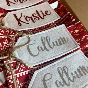 Könnte beinhalten: Vier weiße Geschenkanhänger aus Filz mit spitzem Ende, befestigt an rotem Stoff mit weißem und gelbem nordischem Muster. Zwei Anhänger tragen den Namen Kirstie in dunkelroter Schreibschrift, zwei den Namen Callum in silberner Glitzerschrift.