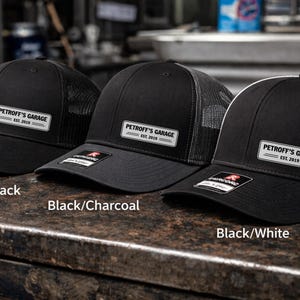 May include: Three black trucker caps with mesh backs. Each cap has a rectangular patch with the text "PETROFF'S GARAGE EST. 2019". The caps are labelled "Black/Black", "Black/Charcoal", and "Black/White".