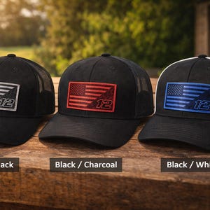 Puede incluir: Se muestran tres gorras de camionero negras con la parte trasera de malla. Cada gorra presenta un parche rectangular con un diseño de bandera estadounidense y el número 12. Las banderas son plateadas, rojas y azules, con los colores de la gorra etiquetados como Negro/Negro, Negro/Carboncillo y Negro/Blanco.