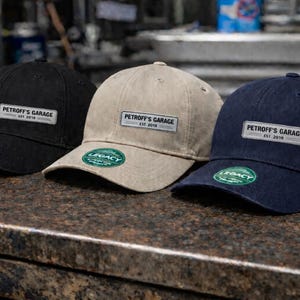 Peut inclure: Trois casquettes de baseball noires, beiges et bleu marine sont présentées sur un comptoir sombre. Chaque casquette porte une étiquette rectangulaire avec le texte "Petroff's Garage EST. 2019". Un autocollant vert "Legacy" est sur la visière.