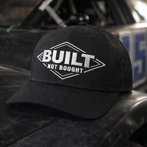 May include: Black trucker hat with the text "BUILT NOT BOUGHT" in white, inside a white diamond shape. The hat has a mesh back and a curved brim. The hat is sitting on a dark surface, with a race car in the background.