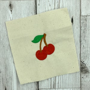 May include: Embroidered design of two red cherries with green stems and leaves on a white fabric background.
