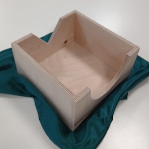 Könnte beinhalten: Eine helle, unfertige Holzkiste mit einem einzigartigen Ausschnitt-Design. Die Kiste ist rechteckig mit einer gebogenen Seite und einem kleineren, abgewinkelten Abschnitt. Sie liegt auf einem dunkelgrünen Satin-Stoff, was einen Kontrast in Texturen und Farben erzeugt.