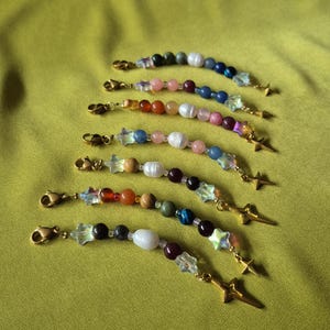 Op de afbeelding: Zeven kralen tasbedels met goudkleurige karabijnsluitingen en stervormige hangers. Elke bedel is samengesteld uit diverse kleurrijke ronde kralen, waaronder witte parels, doorschijnende iriserende sterren en gladde stenen in blauw, rood, groen en bruin.