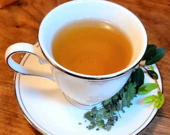 May include: A white teacup and saucer with a gold trim, filled with light brown tea. Green tea leaves are scattered on the saucer.