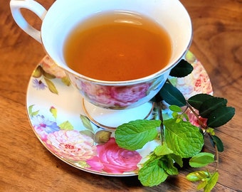 May include: A teacup filled with tea sits on a floral saucer, with a sprig of mint. The teacup and saucer have a white background with pink, red, and purple roses. The tea is a light brown color.